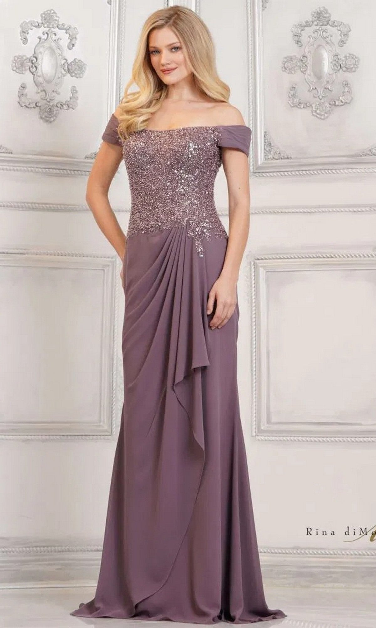A woman wearing the Rina Di Montella RD3128, a long formal mauve chiffon sheath with an off-the-shoulder beaded bodice, stands against an ornate white wall.